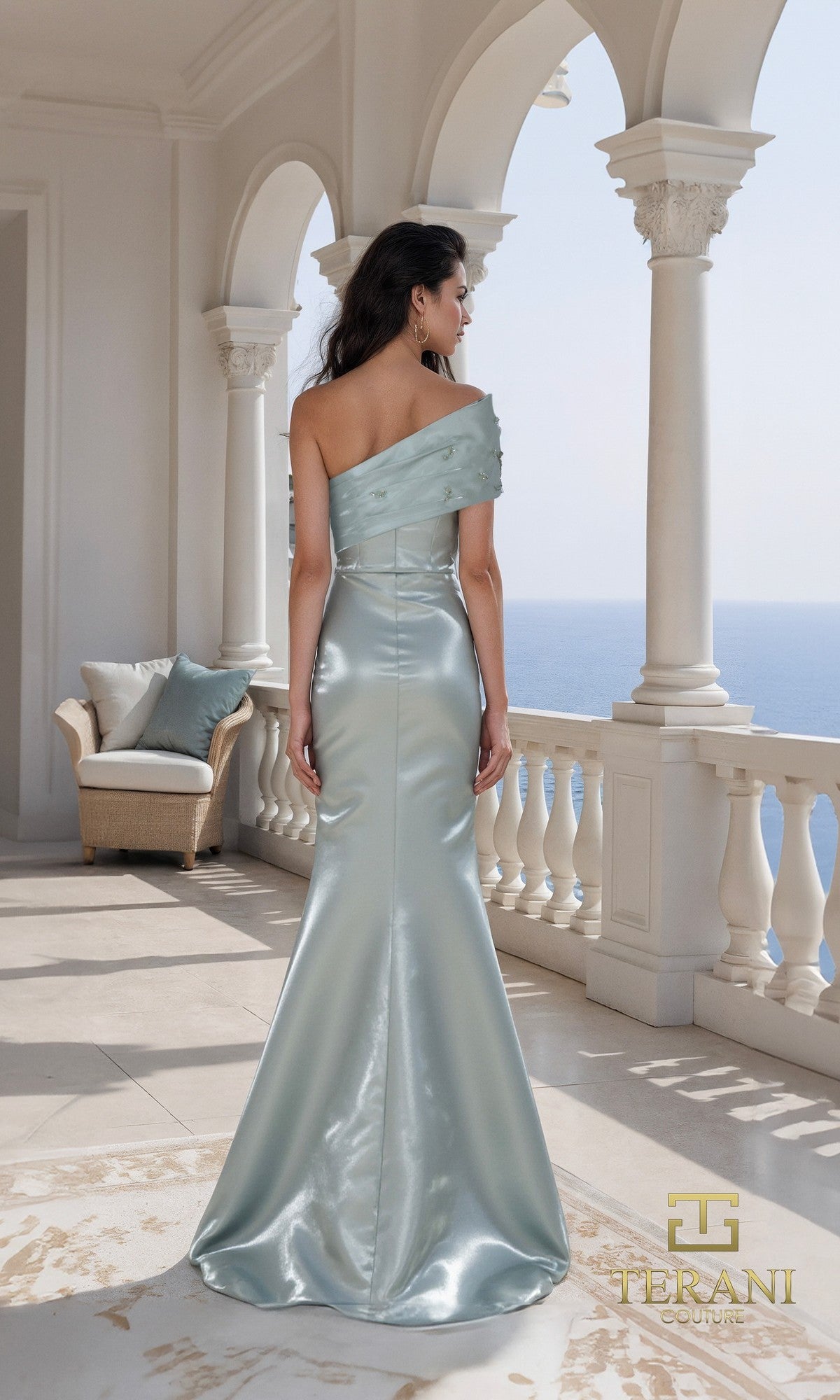 A woman stands on a balcony overlooking the sea, wearing the Long Formal Dress: Terani 261E6227, a light blue one-shoulder gown with a fitted bodice and flared skirt.